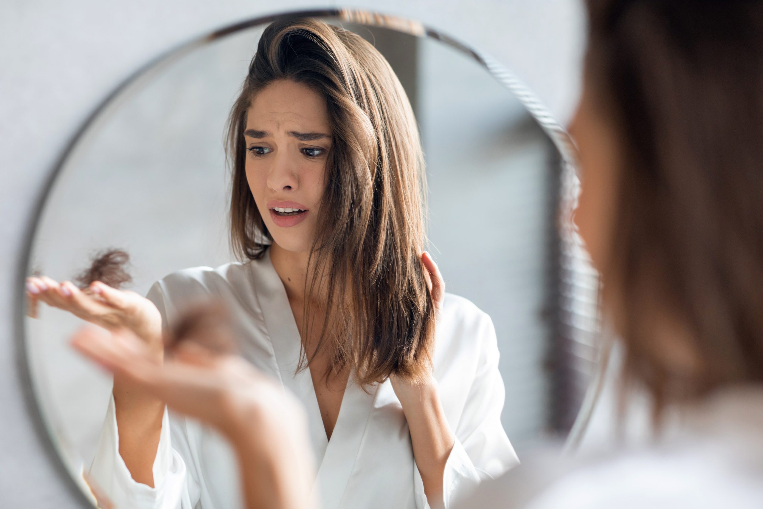 Hairloss Concept. Worried Young Woman Holding Bunch Of Fallen Hair In Hand While Standing Near Mirror In Bathroom, Stressed Beautiful Lady Suffering Alopecia Or Health Problems, Selective Focus