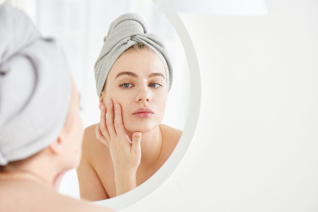 Portrait of  young girl with  towel on head in white bathroom looks and touches her face in the mirror and enjoys youth and hydration. Natural beauty, home care for problem skin