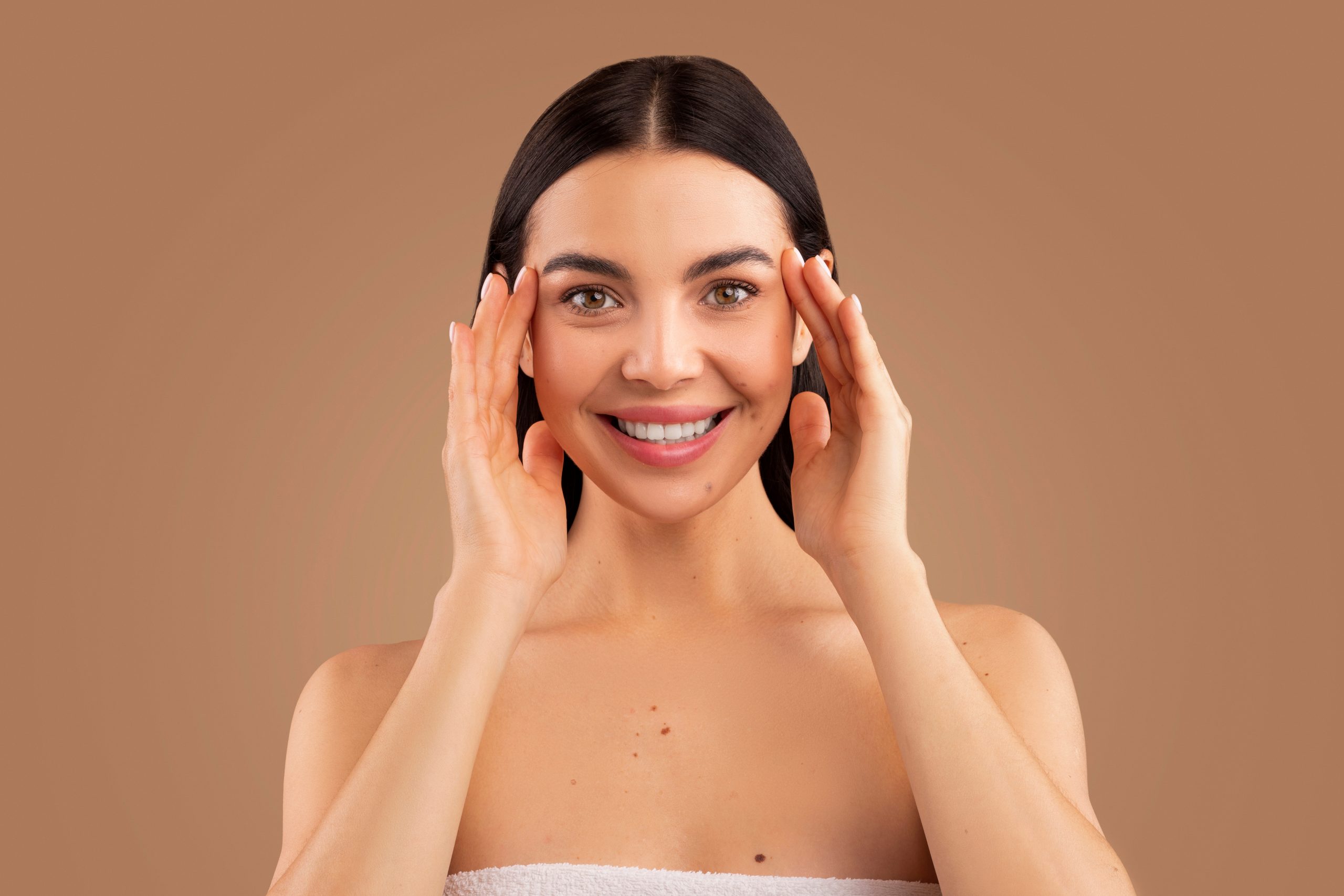 Headshot of beautiful young long-haired brunette half-naked lady rubbing her face and smiling at camera over beige studio background, lady touching silky glowing eye skin, face care concept