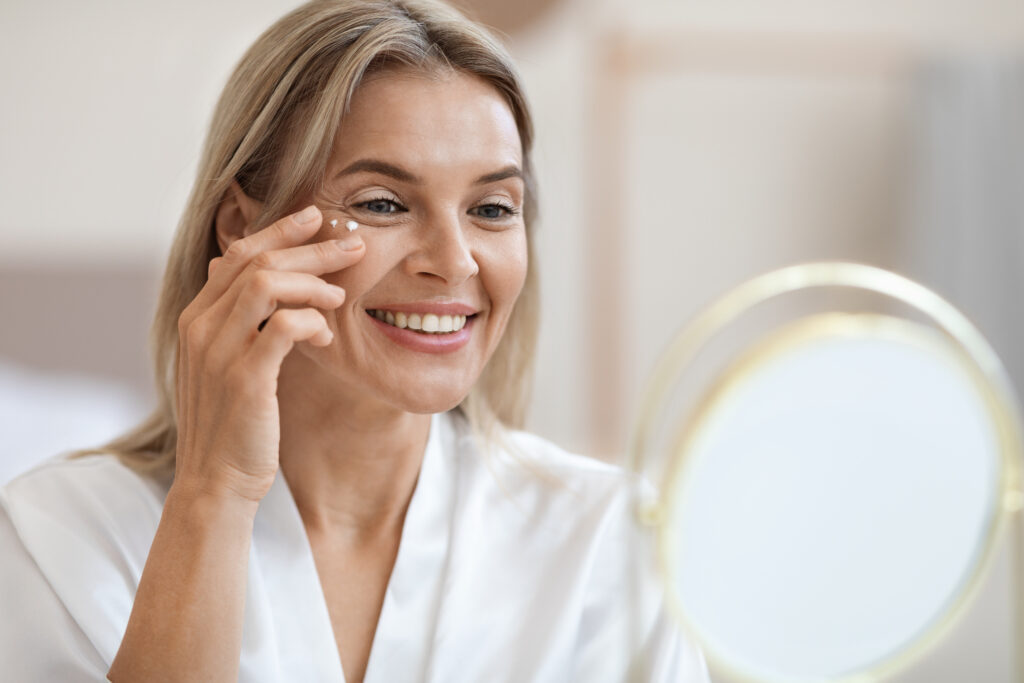Closeup of cheerful beautiful blonde middle aged lady in silky bathrobe applying moisturizing eye cream, looking at mirror and smiling, enjoying face care routine, copy space