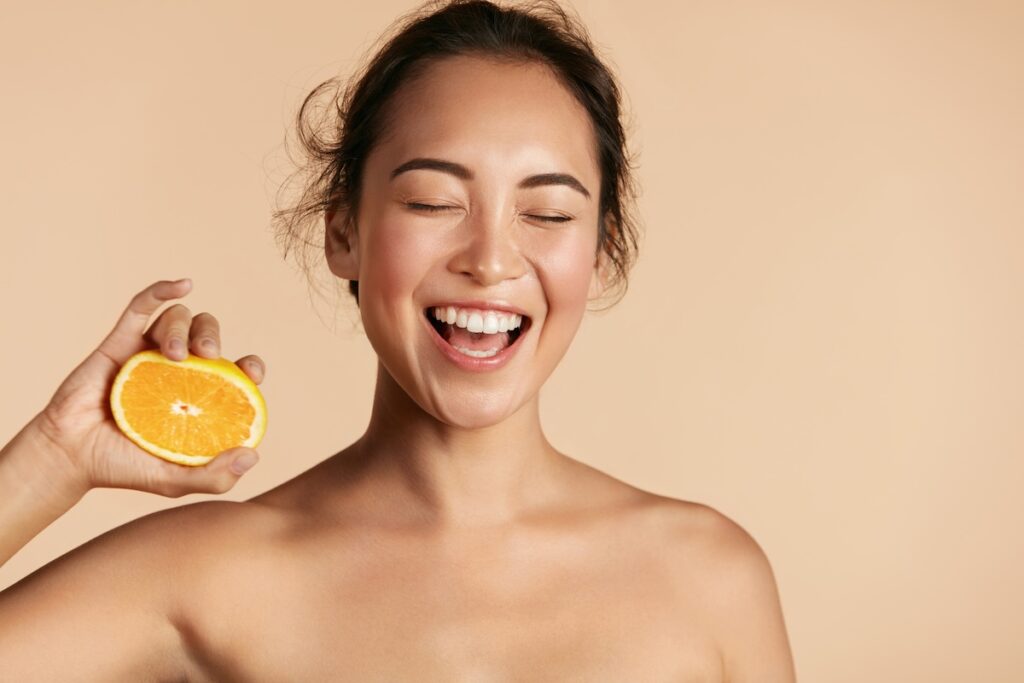 Woman smiling while holding an orange slice, representing how vitamins like Vitamin C contribute to a bright, healthy complexion.