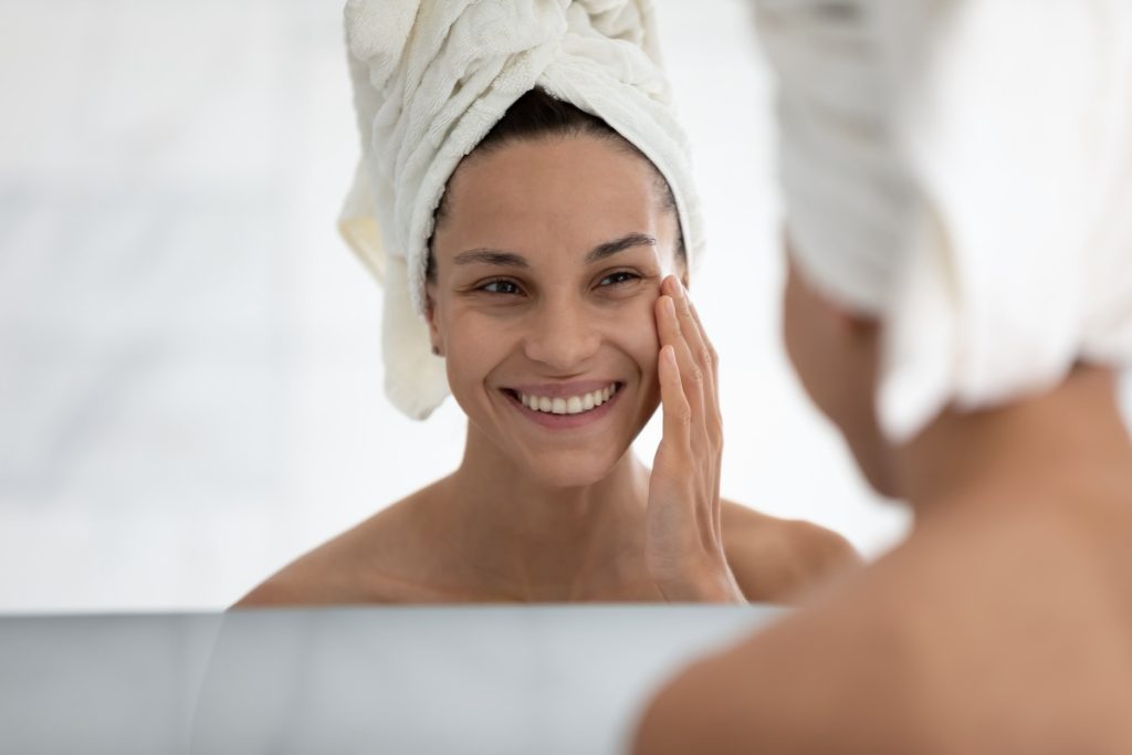 Smiling woman with towel-wrapped hair applying skincare in front of a mirror