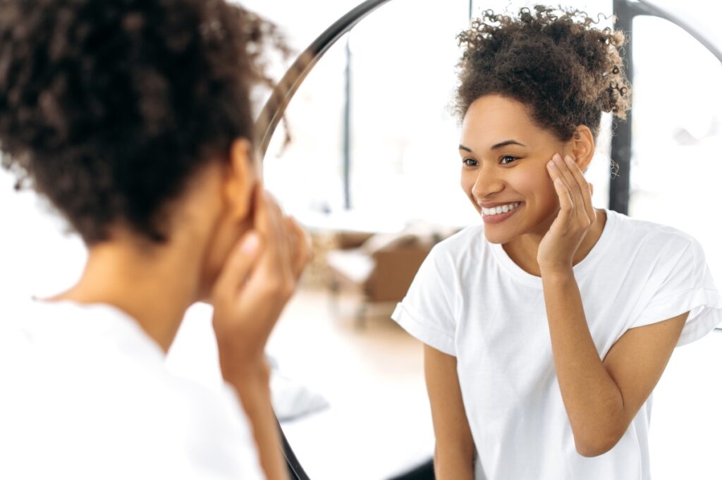 Lovely happy African American curly-haired girl in a white basic t-shirt, stands in front of a mirror in the morning, admires herself, smiles, starts the day well, good mood, looks at her face