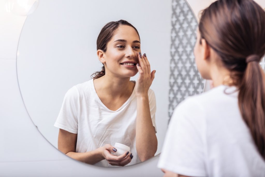 Woman applying moisturizer in front of mirror, practicing a winter skincare routine for hydration.