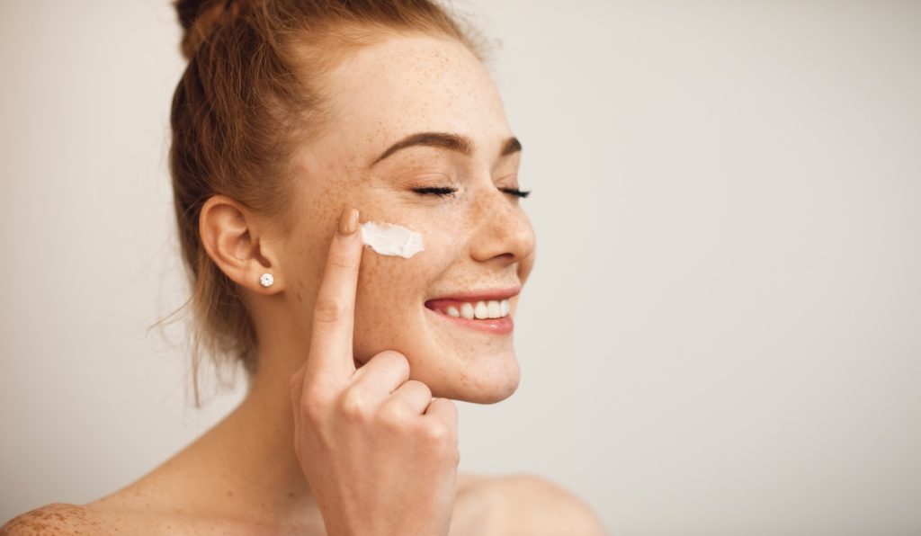Close up of a young female with red hair and freckles applying white cream on her face laughing with closed eyes isolated on white background. Close up of a young woman applying skin cream to her face
