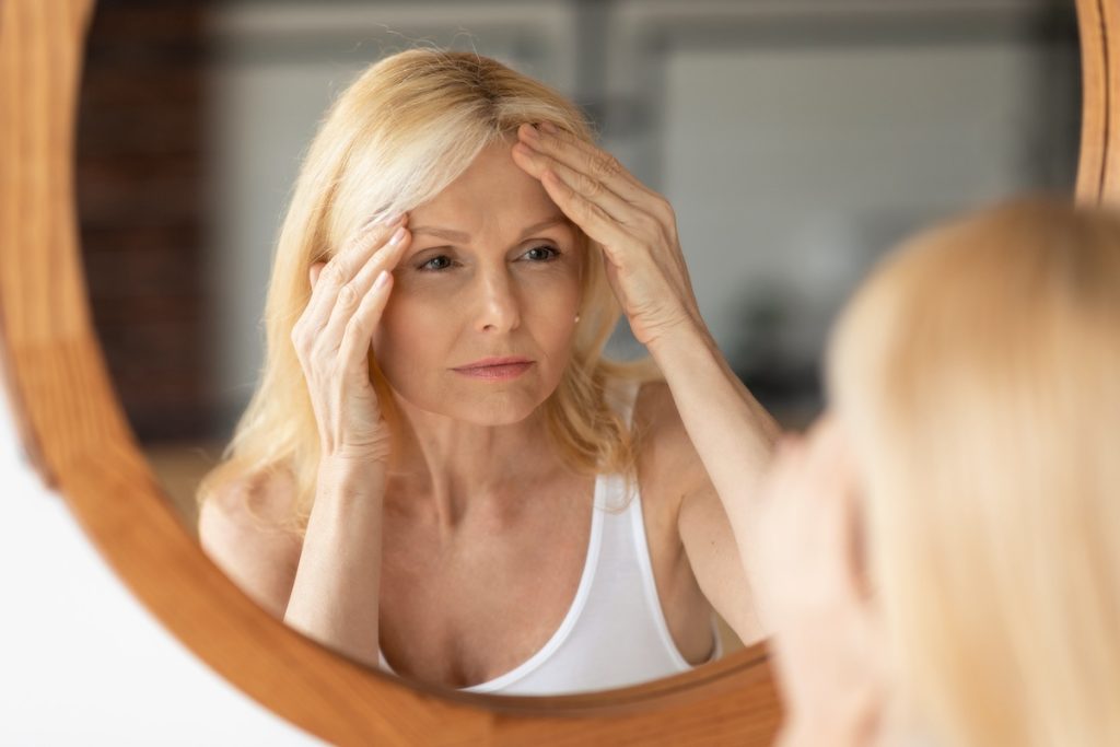 Senior woman looking at mirror and touching forehead, upset aged lady examining fine lines on her face, suffering skin aging, selective focus on reflection